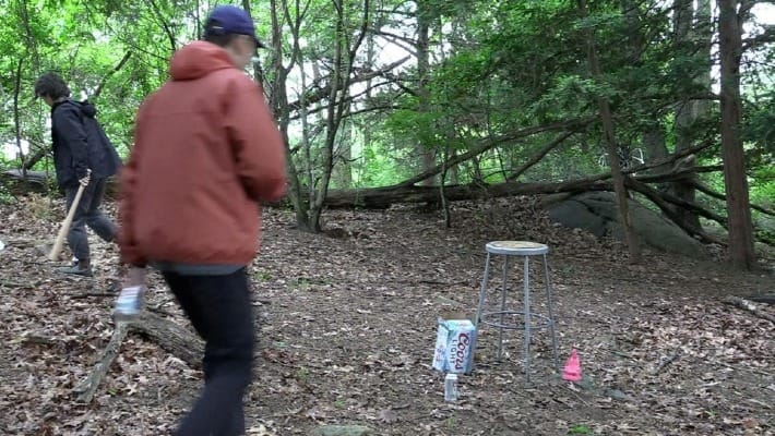 two people are in a forest, one holds a baseball bat; there is also a stool and some Coors