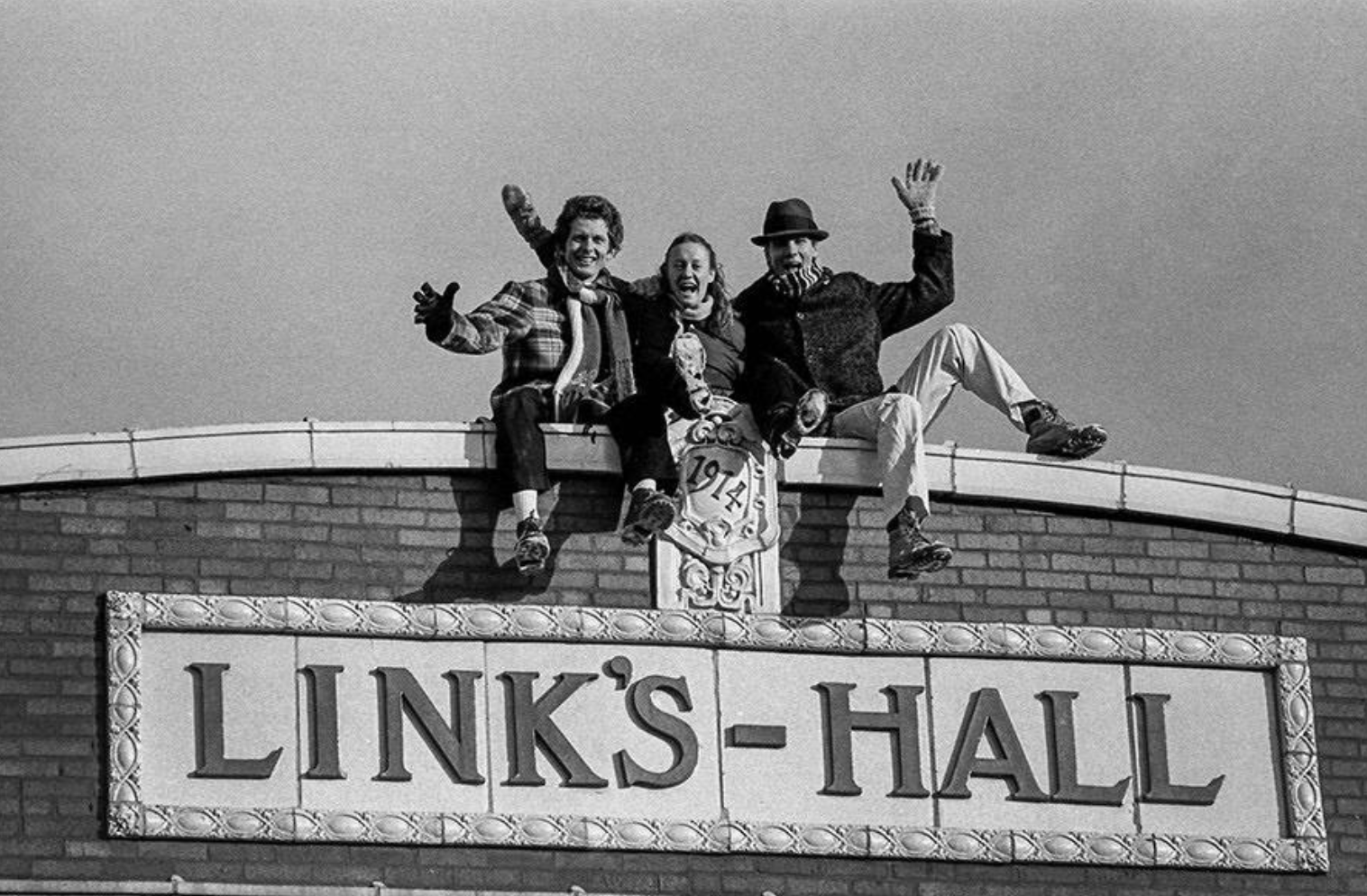 three people perch on the roof of a building that says Link's Hall, limbs out in wacky poses